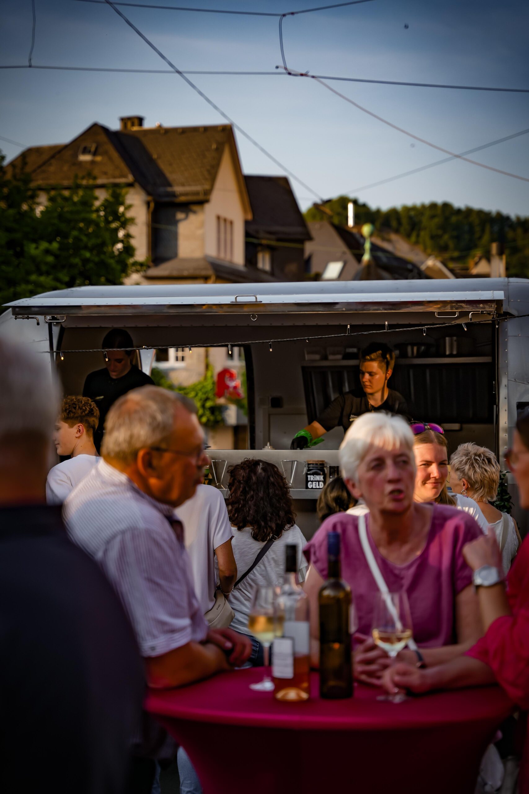 Gäste mit Weingläsern an einem rot eingedeckten Stehtisch vor dem Foodtruck