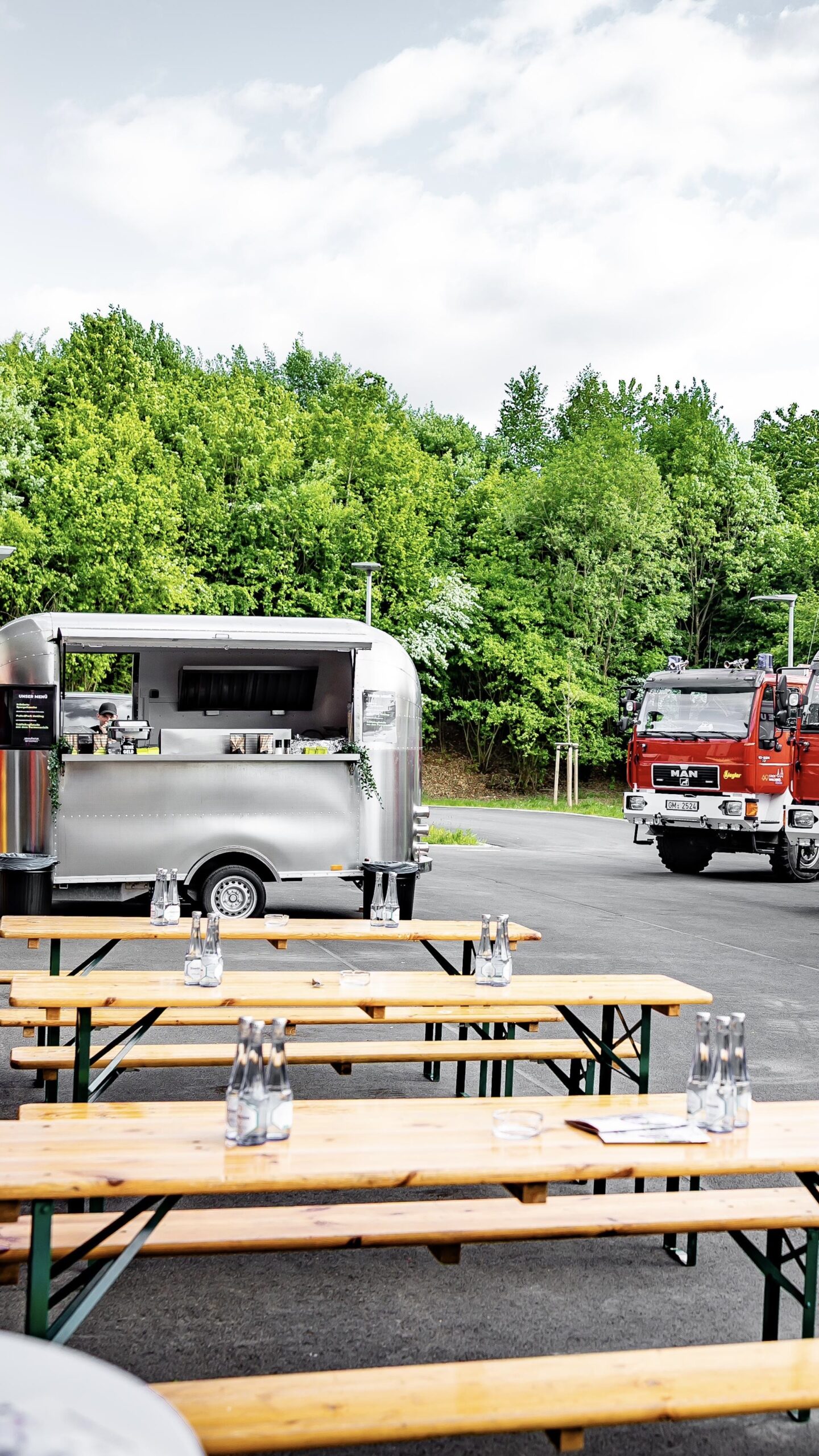 Airstream-Foodtruck mit Bierzeltgarnituren auf einem Platz im Grünen, daneben rote Feuerwehrfahrzeuge