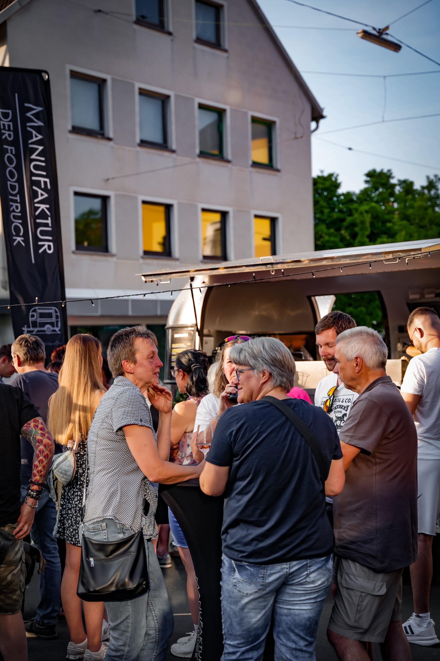 Gäste an Stehtischen vor dem Foodtruck mit ManuFaktur-Beachflag in der Abendsonne