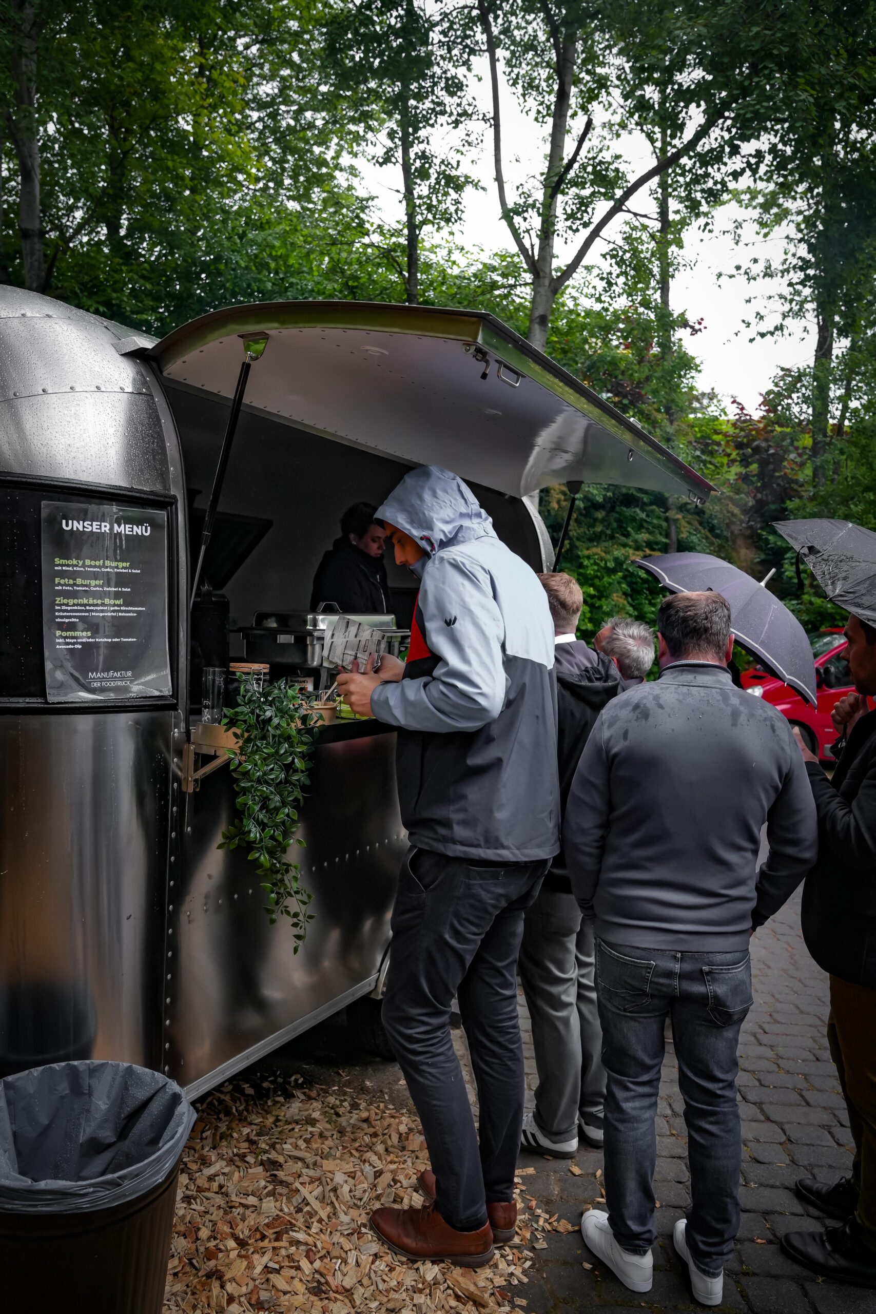 Gäste in Regenjacken und mit Schirmen warten am Foodtruck mit Menütafel
